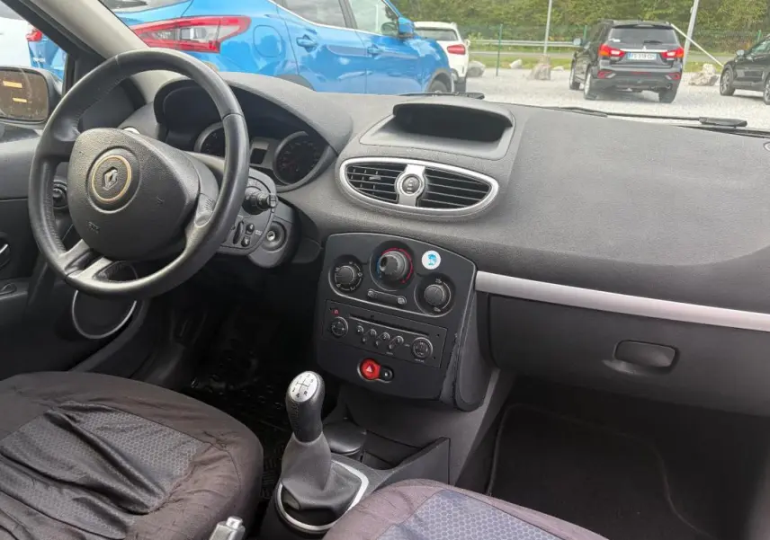 Vue intérieure côté conducteur de la Renault Clio III gris 2005, avec tableau de bord et levier de vitesses manuel visibles.