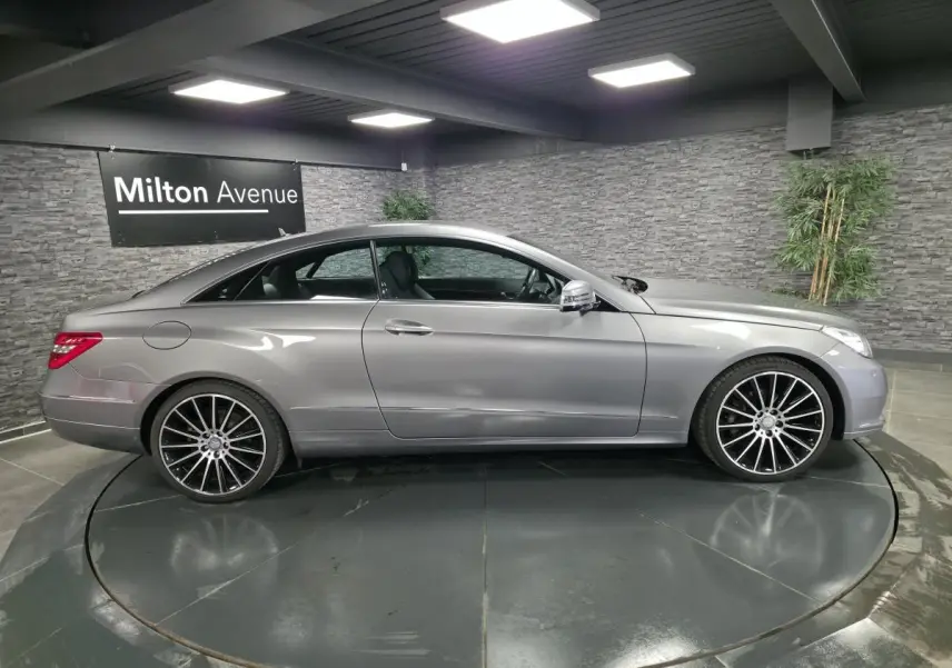 Profil côté gauche d'une Mercedes Classe E Coupé E 200 gris métallisé avec jantes alliage 19 pouces en intérieur showroom.