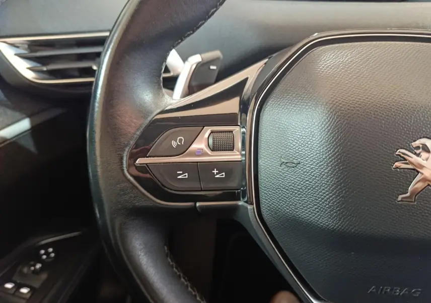 Gros plan sur le volant cuir noir côté gauche du Peugeot 3008 blanc, avec commandes audio et logo lion visible.