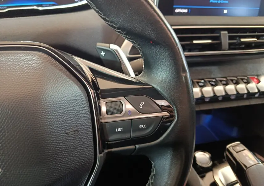 Gros plan sur le volant cuir noir du Peugeot 3008 blanc, avec commandes audio et écran tactile en arrière-plan.