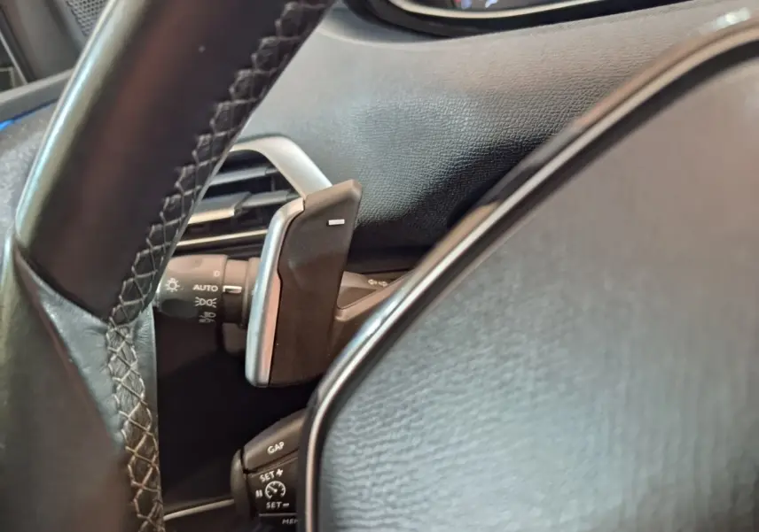 Gros plan sur le volant cuir noir côté gauche du Peugeot 3008 blanc, avec commandes au volant et palettes de changement de vitesse.