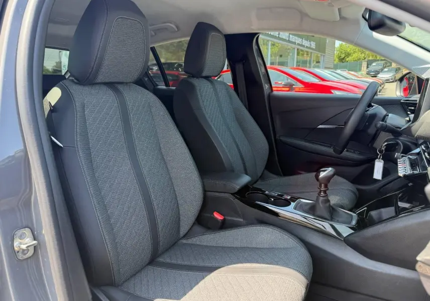 Vue intérieure côté conducteur de la Peugeot 208 gris Artense, sièges tissu gris et levier de vitesse manuel visible.