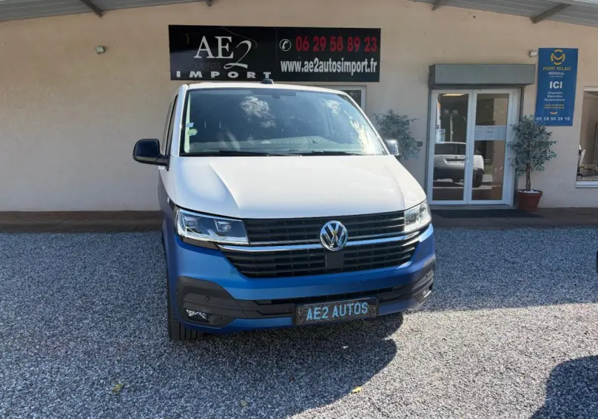 Vue avant d'un Volkswagen Transporter blanc avec pare-chocs bleu, stationné devant un bâtiment commercial.