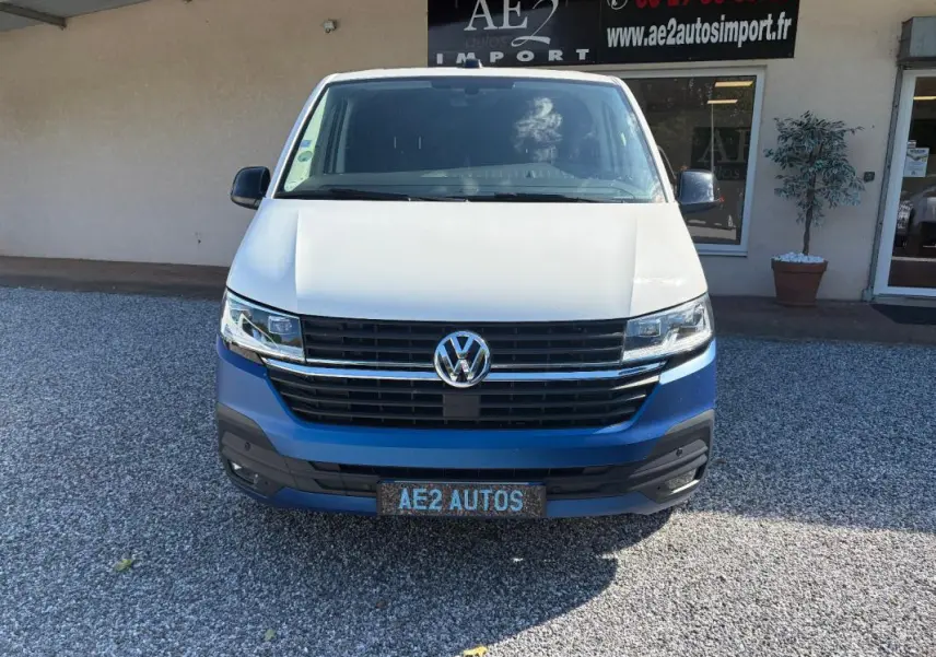 Vue avant du Volkswagen Transporter blanc avec calandre noire et logo VW bien visible, stationné devant un bâtiment.