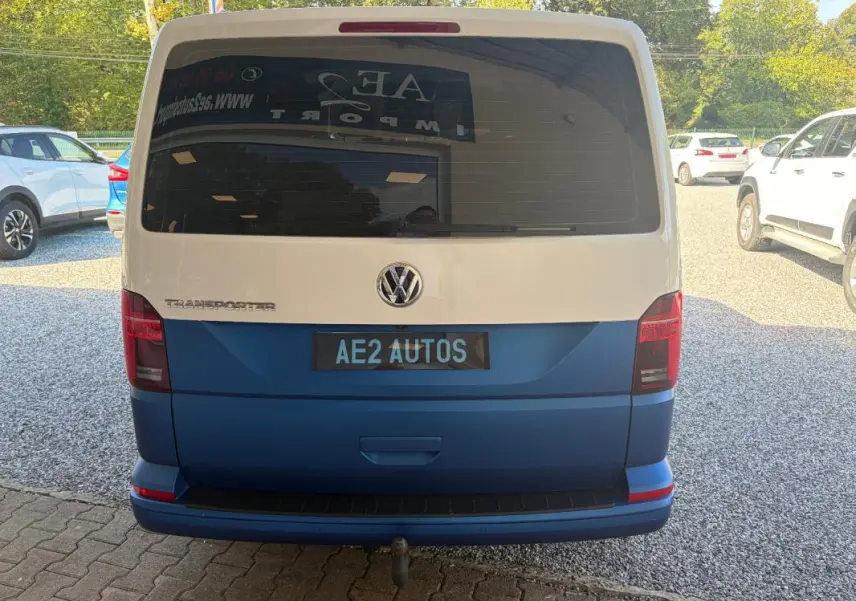 Vue arrière d’un Volkswagen Transporter blanc et bleu, avec vitre arrière teintée et plaque AE2 AUTOS visible.