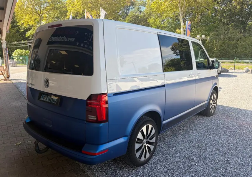 Vue 3/4 arrière droite d’un Volkswagen Transporter 2.0 TDI blanc et bleu avec jantes aluminium et vitres teintées.
