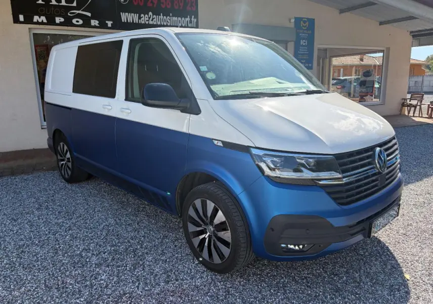 Vue 3/4 avant droit d’un Volkswagen Transporter blanc et bleu avec jantes aluminium, stationné devant un bâtiment.