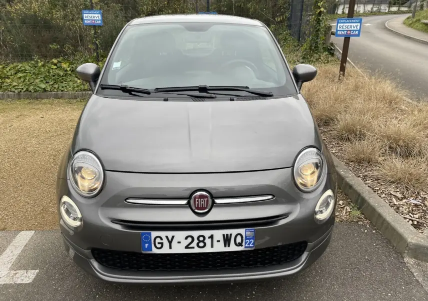 Vue avant d'une FIAT 500 gris foncé avec phares allumés, stationnée sur un parking réservé Rent A Car.