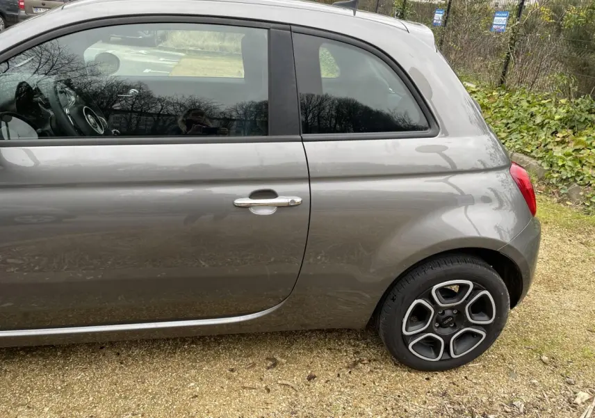 Vue de profil côté gauche d'une FIAT 500 gris foncé avec jante alliage distinctive, stationnée sur sol gravillonné.