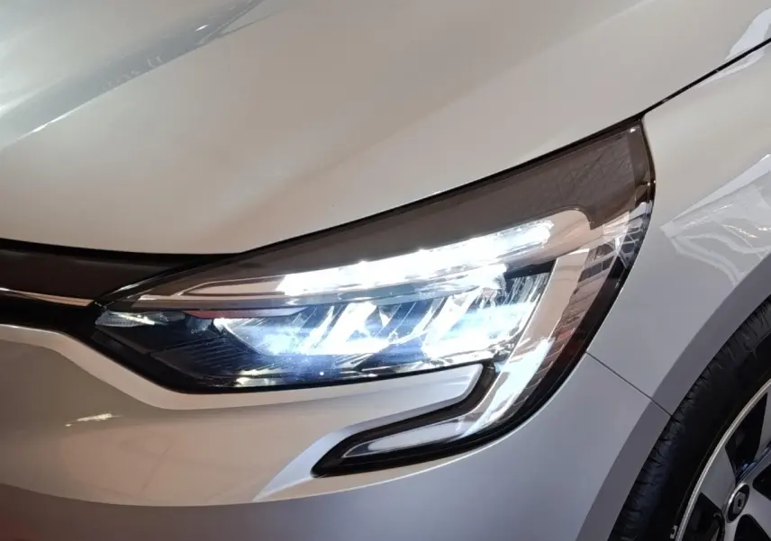 Gros plan sur le phare avant droit allumé d'une Renault Clio gris clair 2023, avec partie de la roue visible.