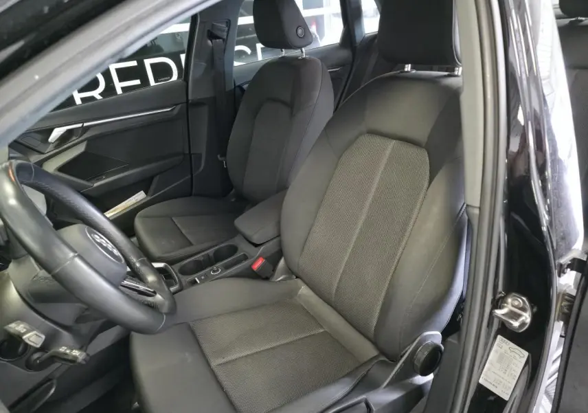 Vue intérieure côté conducteur de l'Audi A3 Sportback 2023 noire, montrant les sièges tissu et le volant cuir multifonctions.