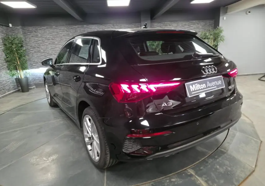 Audi A3 Sportback noir vue 3/4 arrière droit avec feux arrière LED allumés en intérieur showroom.