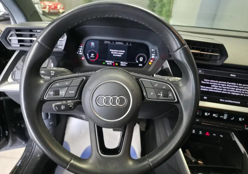 Vue intérieure centrée sur le volant cuir noir multifonctions de l'Audi A3 Sportback 2023 avec tableau de bord digital.