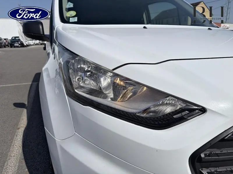 Gros plan sur le phare avant gauche du Ford Transit Connect blanc glacier, montrant les détails du bloc optique et la calandre partielle.