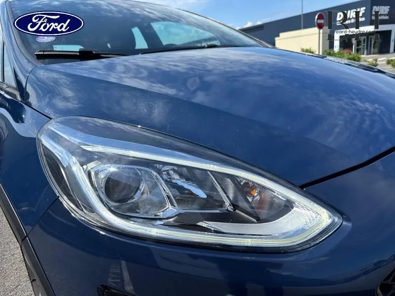 Gros plan sur le phare avant gauche d'une Ford Fiesta 2018 bleu Abysse, avec capot et pare-brise partiellement visibles.