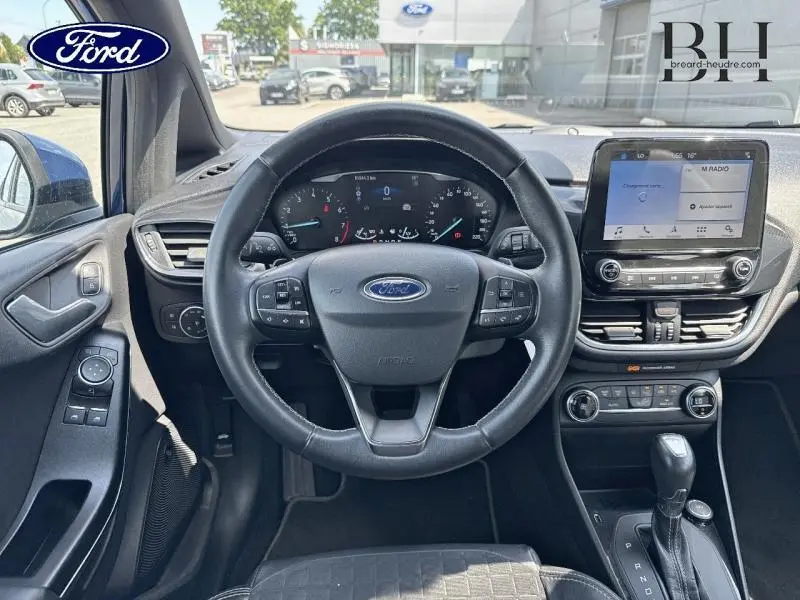 Vue intérieure centrée sur le volant cuir et tableau de bord de la Ford Fiesta Bleu Abysse avec écran tactile et boîte automatique.
