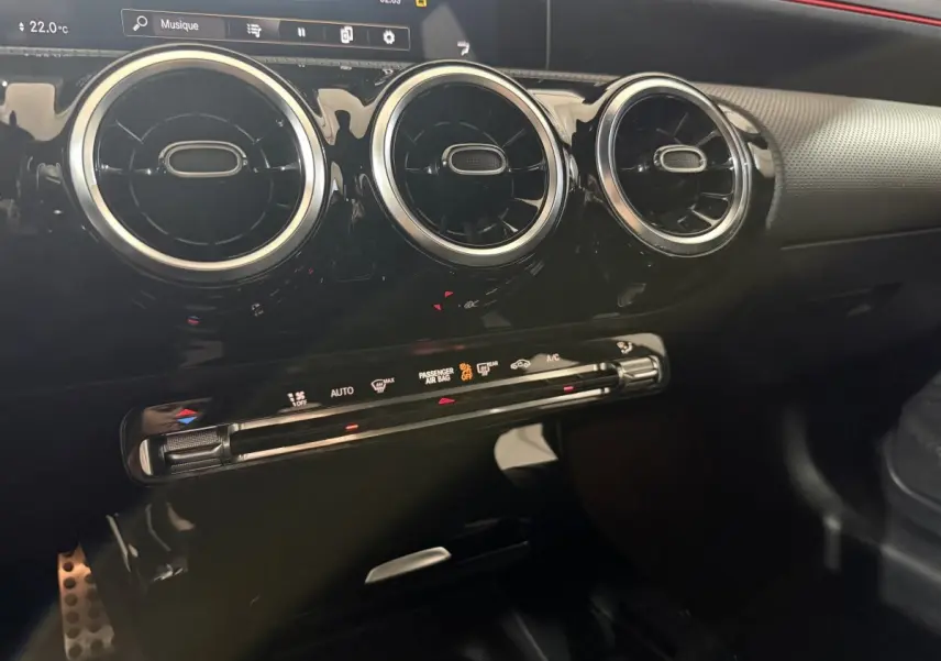 Gros plan intérieur sur la console centrale noire brillante avec aérateurs ronds chromés et bandeau tactile rouge de la Mercedes Classe A AMG Line.