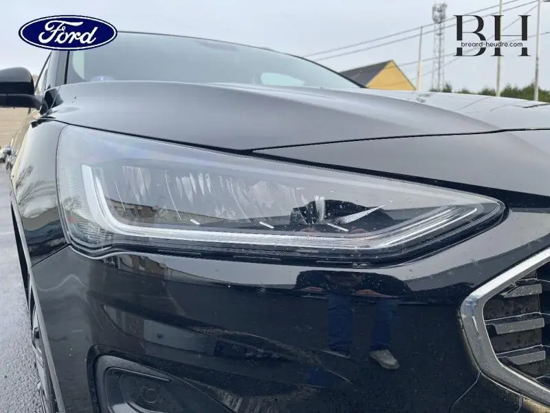 Gros plan sur le phare avant gauche du Ford Focus SW noir Agate métallisé, soulignant la calandre chromée.