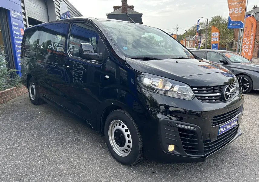 Opel Vivaro Combi noir vu en 3/4 avant droit, stationné devant un garage avec panneaux publicitaires.