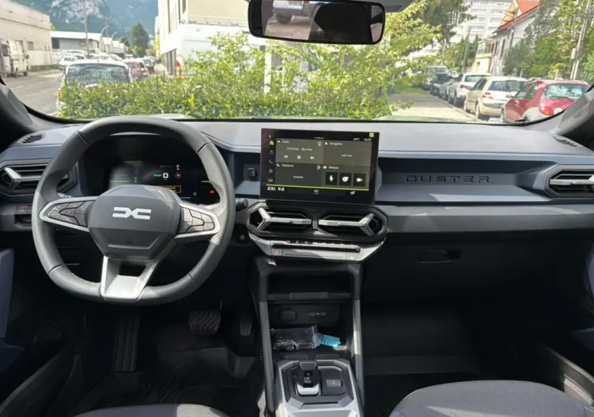 Vue intérieure avant du tableau de bord du Dacia Duster 2025, avec écran tactile central et volant multifonction noir.