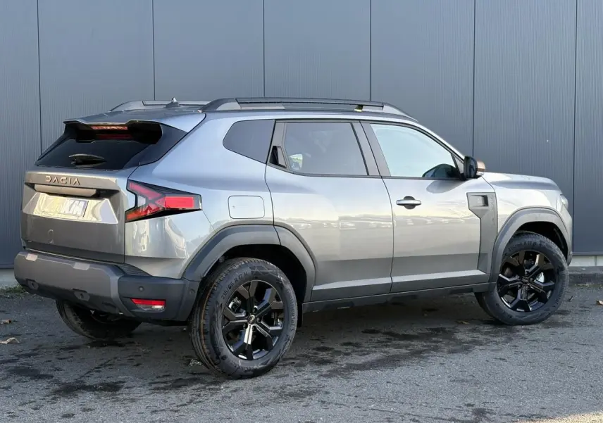 Vue 3/4 arrière droite du Dacia Duster 2025 gris schiste avec jantes noires et barres de toit noires.