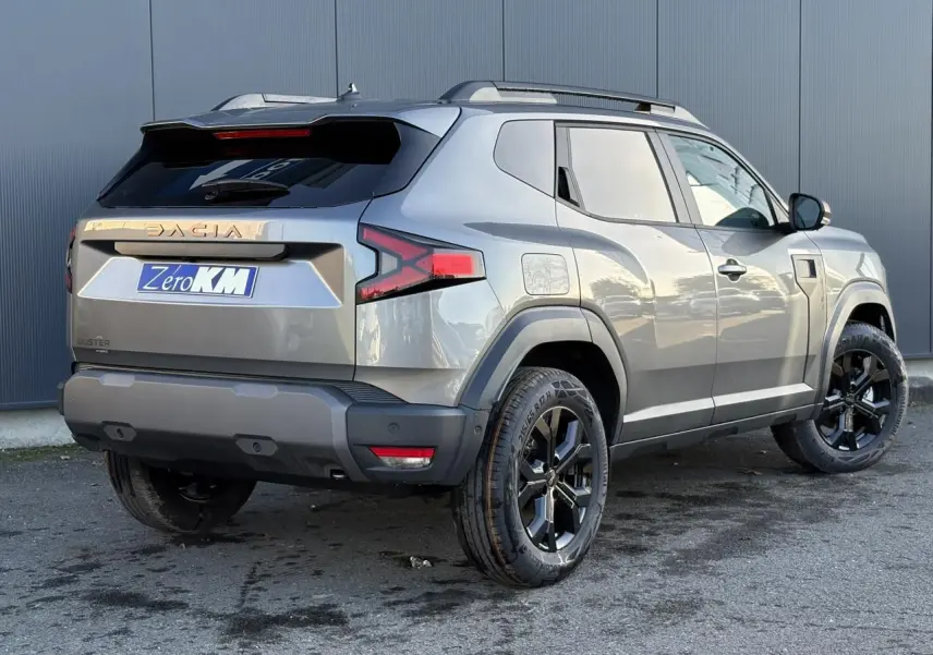 Vue 3/4 arrière droite du Dacia Duster 2025 gris schiste avec jantes noires et barres de toit longitudinales.