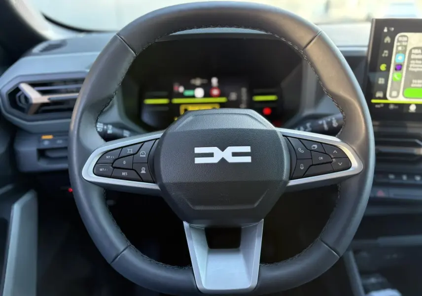 Vue rapprochée du volant noir du Dacia Duster 2025 avec commandes intégrées et tableau de bord digital en arrière-plan.