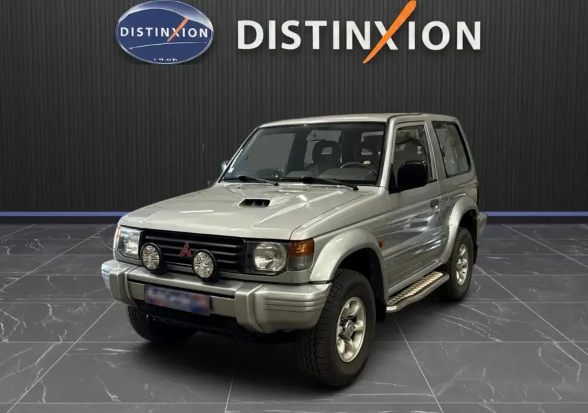 Mitsubishi Pajero Court gris aluminium vu en 3/4 avant droit, avec marchepieds et phares additionnels sur calandre.