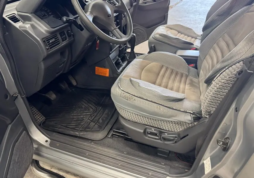 Vue intérieure côté conducteur du Mitsubishi Pajero Court gris aluminium de 1997, avec sièges tissu gris et tapis caoutchouc.