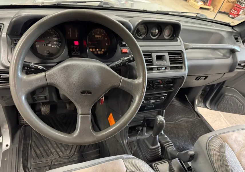 Vue intérieure du tableau de bord gris et volant du Mitsubishi Pajero Court 2.8 TD 1997 avec boîte manuelle et tapis caoutchouc.