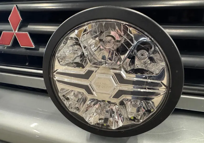 Gros plan sur le phare rond avant du Mitsubishi Pajero Court gris aluminium, avec logo rouge distinctif sur la calandre.