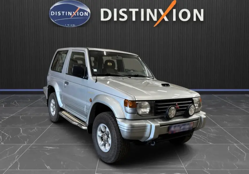 Mitsubishi Pajero Court gris aluminium vu en 3/4 avant droit avec marchepieds et deux phares additionnels frontaux.