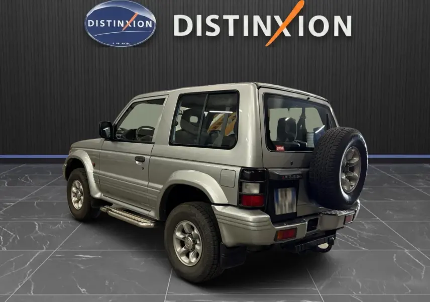 Vue 3/4 arrière droite du Mitsubishi Pajero Court gris aluminium de 1997 avec roue de secours et marchepieds visibles.
