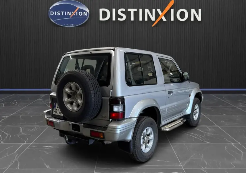 Vue 3/4 arrière droite du Mitsubishi Pajero Court gris aluminium de 1997 avec roue de secours et marchepieds visibles.