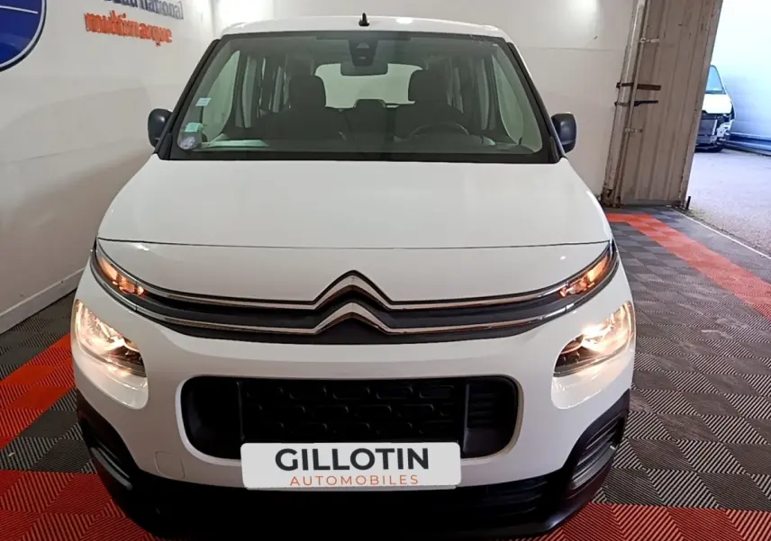 Vue avant d'un Citroën Berlingo XL blanc avec phares allumés, sur sol à damiers rouge et noir en intérieur.