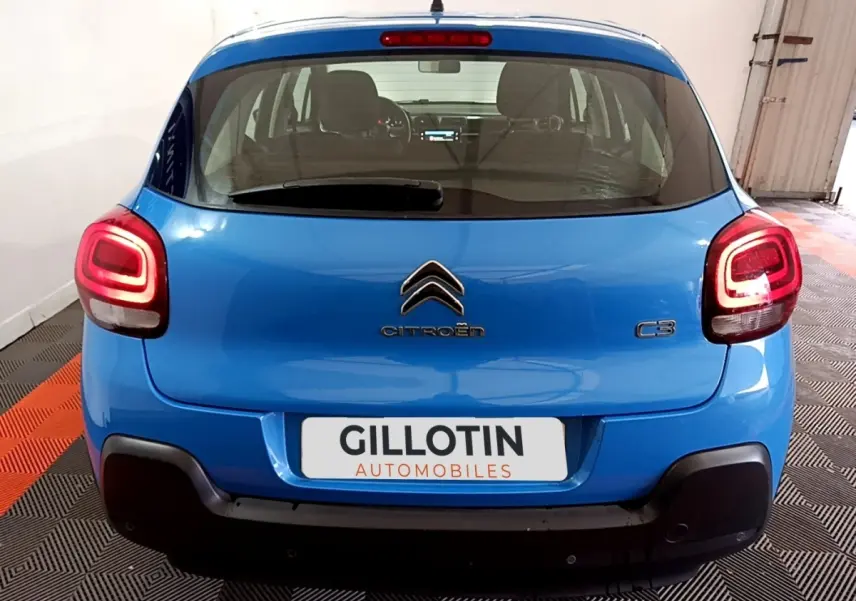 Vue arrière d'une Citroën C3 bleu vif avec feux arrière allumés, stationnée sur un sol à motifs noir et orange.