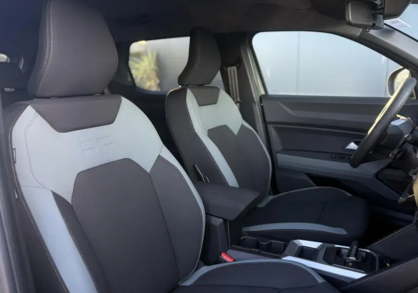 Vue intérieure côté conducteur du Dacia Duster 2025, sièges bi-ton gris et noir avec accoudoir central.