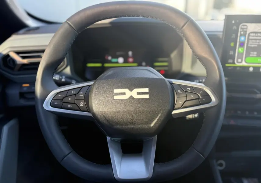 Vue rapprochée du volant noir du Dacia Duster 2025 avec commandes intégrées et tableau de bord digital en arrière-plan.