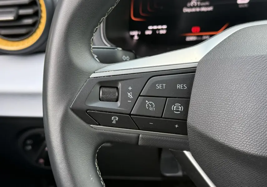 Gros plan sur les commandes gauche du volant noir du SEAT Arona 2025, avec tableau de bord digital en arrière-plan.