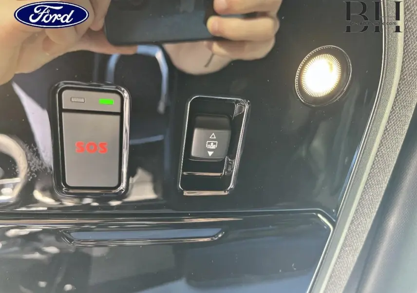 Panneau de commandes intérieur noir brillant avec bouton SOS rouge et éclairage, reflet de mains tenant un téléphone.