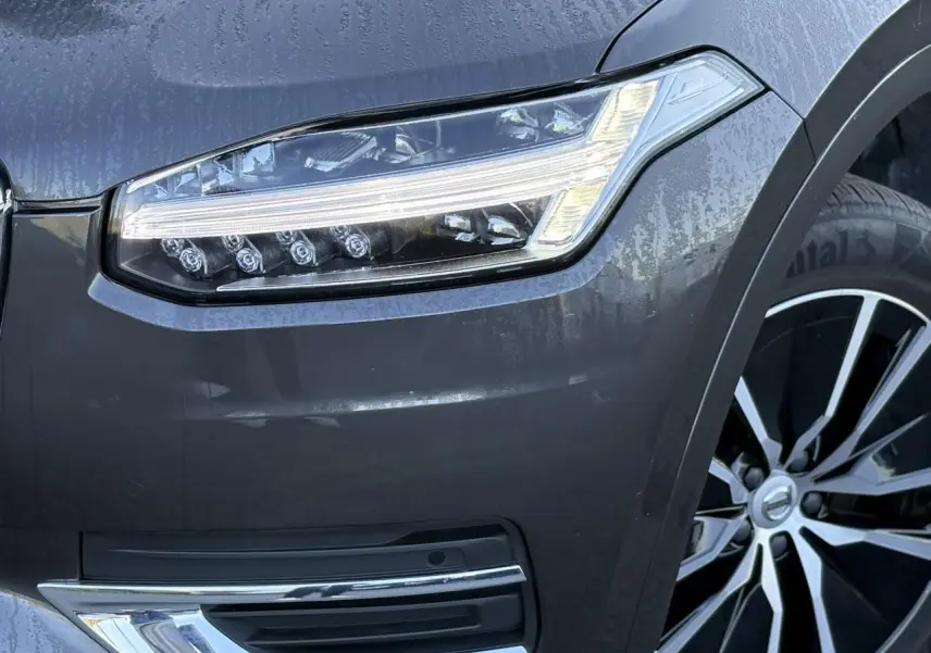 Gros plan sur le phare avant gauche et la jante alliage 19" noir diamant du Volvo XC90 gris Platinium 2024.