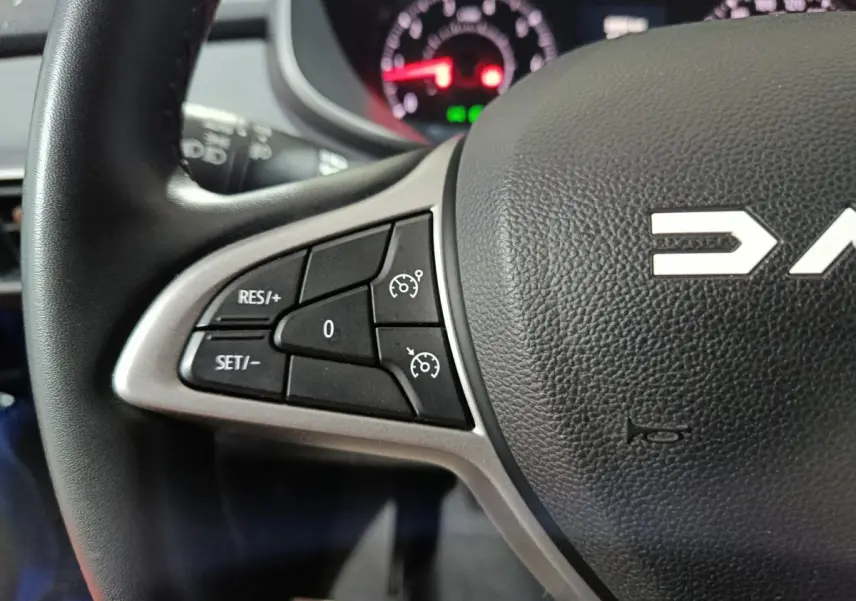 Gros plan sur le volant noir de la Dacia Sandero 2024, avec commandes de régulateur de vitesse à gauche et tableau de bord flou en arrière-plan.