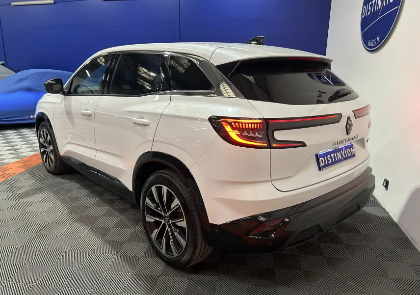 Vue 3/4 arrière droite du Renault Austral blanc nacré avec feux arrière allumés et hayon motorisé visible en intérieur showroom.