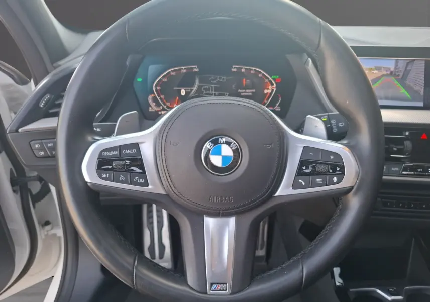Vue intérieure centrée sur le volant cuir M Sport d'une BMW Série 1 blanche avec tableau de bord numérique et caméra de recul.