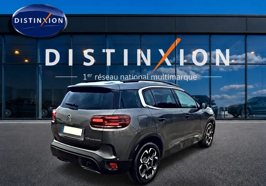 Vue 3/4 arrière droite d'un Citroën C5 Aircross gris 2025 avec barres de toit et jantes alu devant un showroom Distinxion.