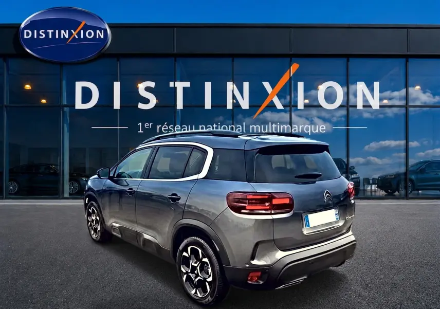 Vue 3/4 arrière droite d'un Citroën C5 Aircross gris 2025 avec jantes alu et barres de toit devant un showroom Distinxion.