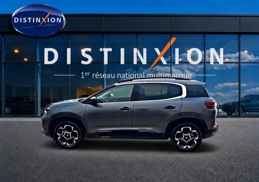 Profil côté gauche du Citroën C5 Aircross gris 2025, avec jantes alu et barres de toit visibles devant une vitrine Distinxion.