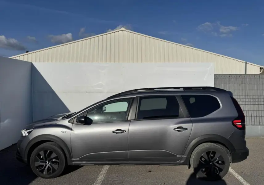 Profil droit du Dacia Jogger gris schiste métallisé avec barres de toit et jantes noires sur parking extérieur.