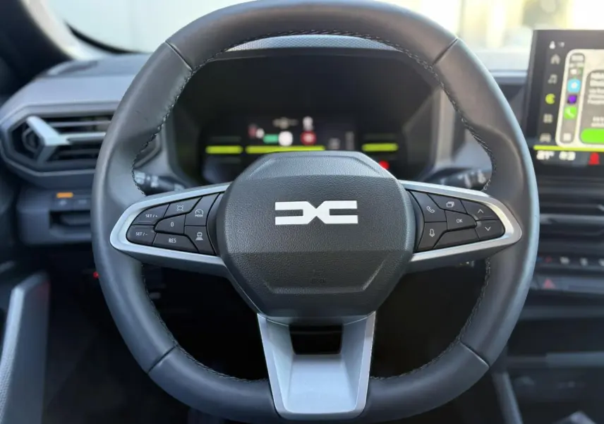 Vue frontale du volant noir du Dacia Duster 2025 avec commandes intégrées et tableau de bord digital en arrière-plan.
