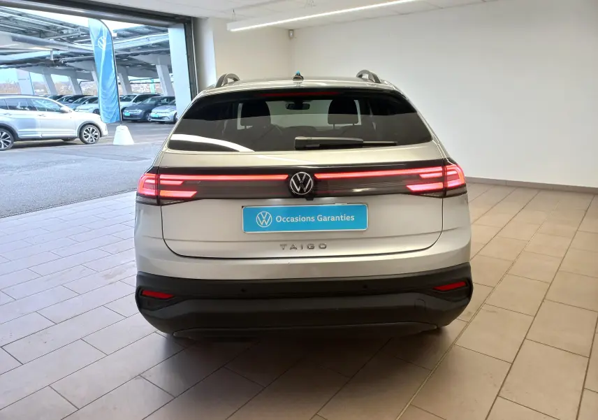Vue arrière d'un Volkswagen Taigo 2025 en peinture métallisée Reflet d'argent avec feux arrière LED allumés dans un showroom.